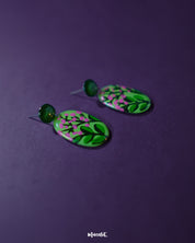 A close-up view of the Kalibaan drop earrings resting on a textured surface, highlighting the vibrant hand-painted floral design in shades of green and pink. The detailing showcases artisanal craftsmanship and colorful statement appeal — Muselot.