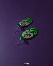 The Kalibaan handmade drop earrings placed against a soft purple background, highlighting the hand-painted floral details and intricate metalwork. The rich contrast showcases the earrings' artistic charm and vibrant elegance — Muselot.