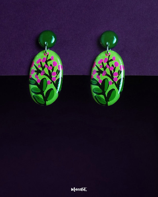 The Kalibaan drop earrings are showcased against a deep purple background, featuring a vivid green oval base hand-painted with delicate pink floral motifs,highlighting the bold charm of nature-inspired design, the earrings are topped with glossy round studs that add a playful yet grounded touch — Muselot.