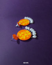 A striking pair of Kesarbaan earrings featuring bold orange and yellow hand-painted patterns, accentuated with dangling white pearls that add a graceful sway – Muselot.