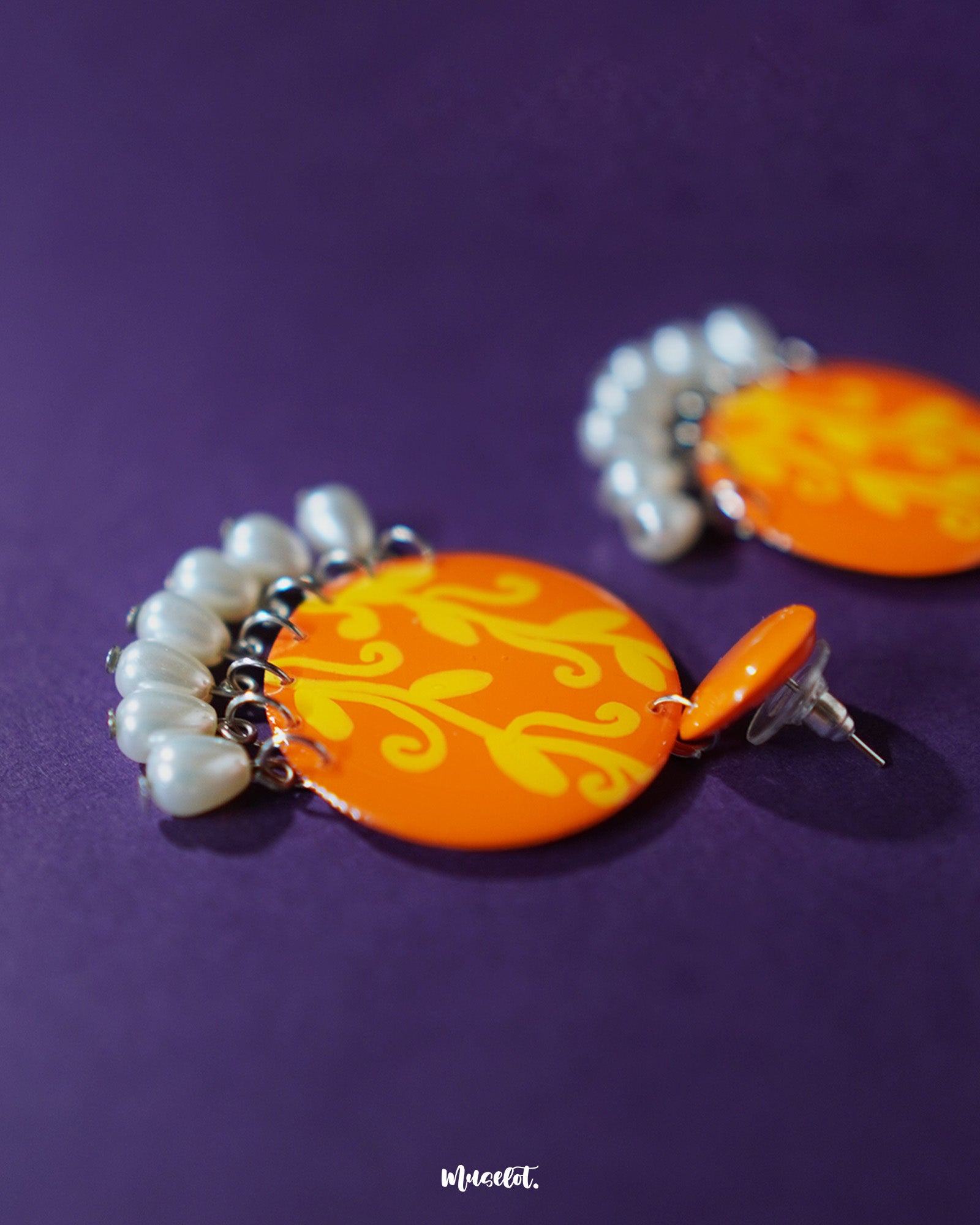 A close-up of the Kesarbaan orange and yellow drop earrings revealing the intricate brushstrokes of the floral design, paired with elegant white pearl drops that add charm to their festive handcrafted appeal — Muselot.
