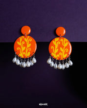 Kesarbaan pearl drop earrings set against a deep purple backdrop, featuring vibrant orange discs adorned with yellow folk motifs and finished with delicate white pearl drops that add a touch of festive elegance – Muselot.