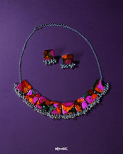 Khushk Gul handmade floral necklace set featuring a silver chain necklace and matching square stud earrings, adorned with vibrant pink, orange, and green abstract floral patterns and delicate silver bead accents, beautifully showcased on a deep purple background – Muselot