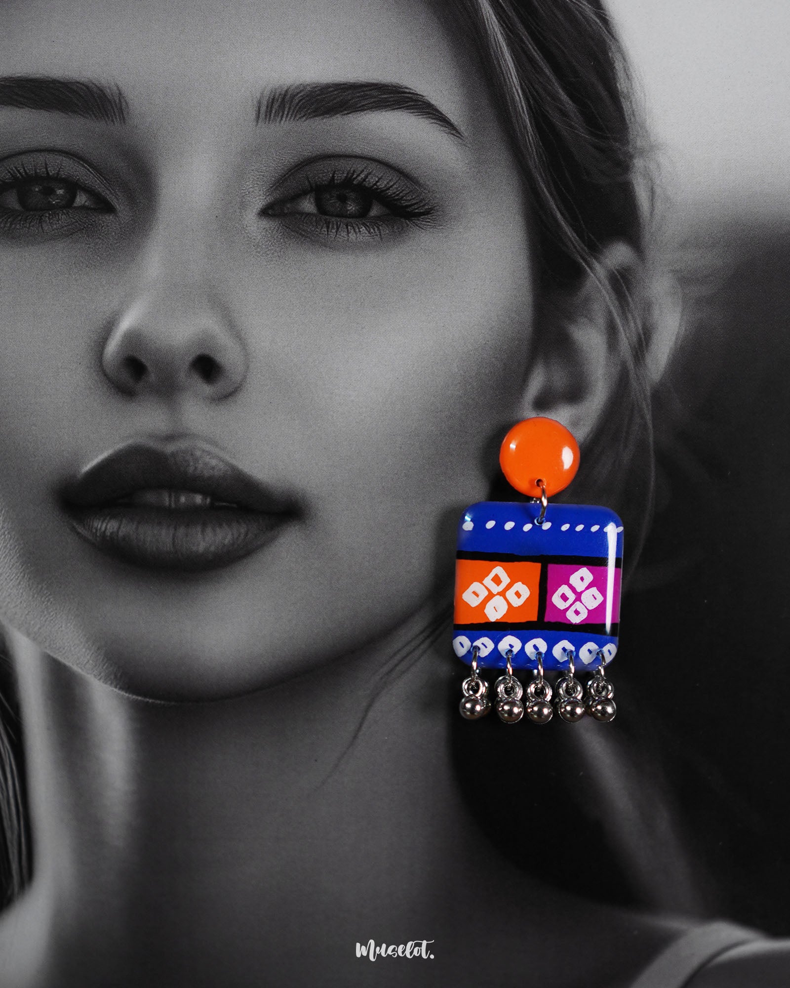 Close-up of model wearing the Leheriya boho earrings, showcasing bright orange round studs connected to blue rectangular drops with orange and magenta blocks, detailed with white geometric patterns and dotted accents, finished with dangling silver beads, capturing their vibrant and artistic design – Muselot.