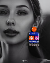 Close-up of model wearing the Leheriya boho earrings, showcasing bright orange round studs connected to blue rectangular drops with orange and magenta blocks, detailed with white geometric patterns and dotted accents, finished with dangling silver beads, capturing their vibrant and artistic design – Muselot.