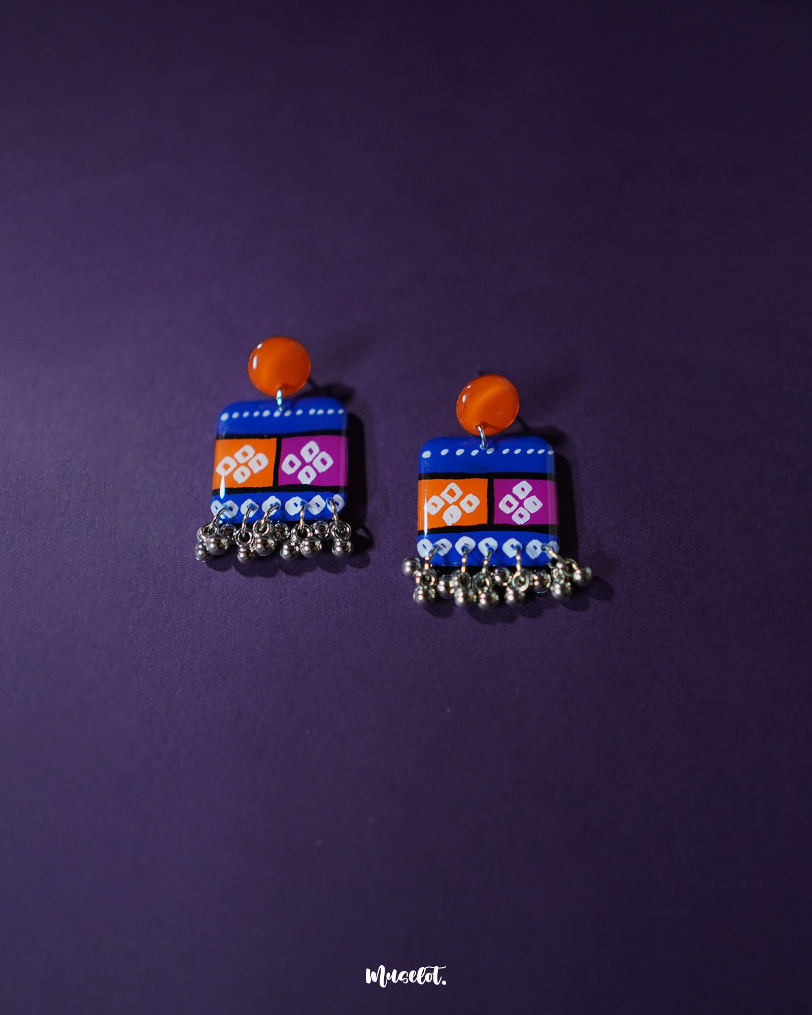 Leheriya traditional art drop earrings displayed on a purple background, featuring bright orange round studs attached to blue rectangular drops with orange and magenta blocks, adorned with white geometric patterns and dotted detailing, finished with dangling silver beads – Muselot.