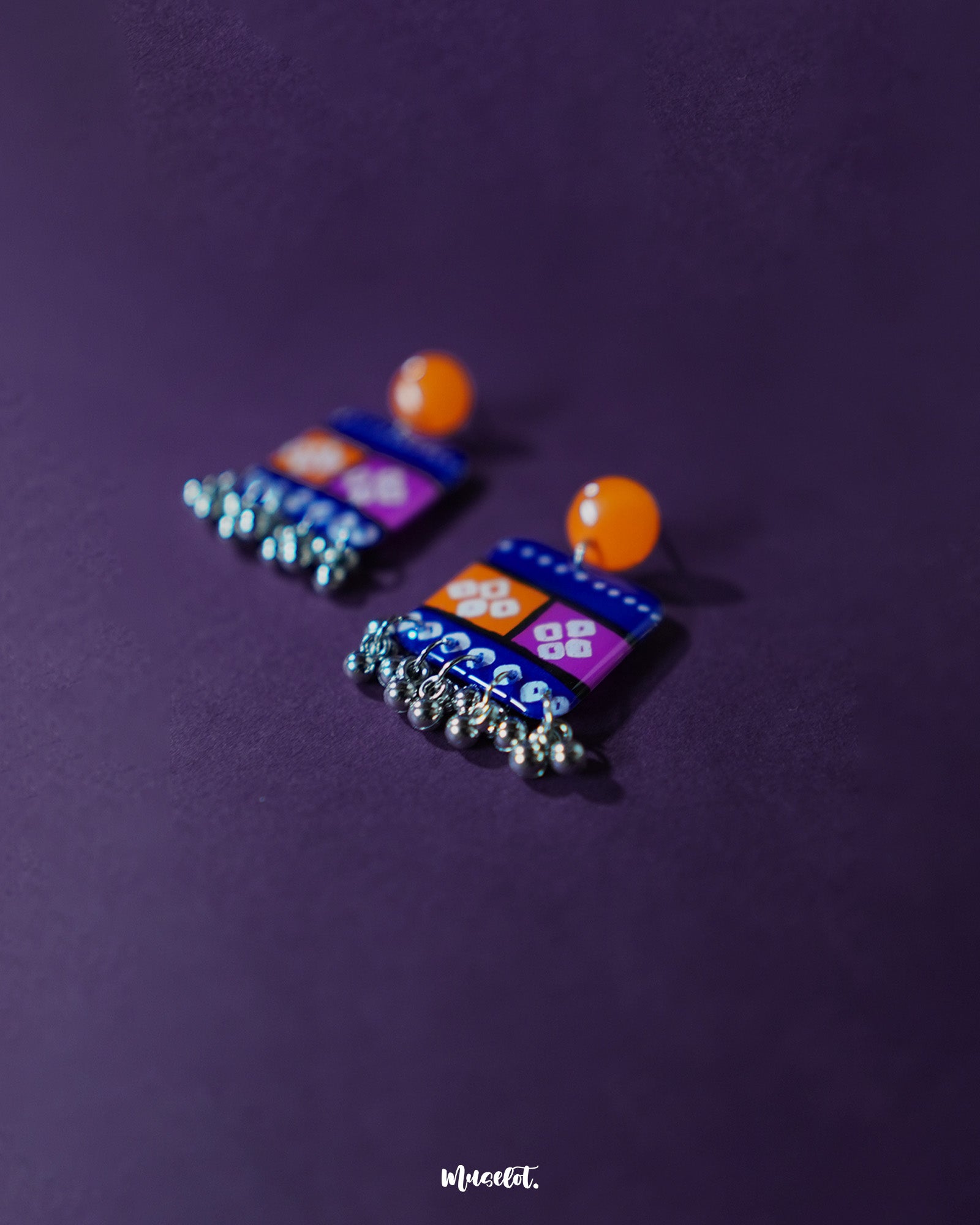 Low-angle view of the Leheriya traditional art drop earrings, showcasing bright orange round studs attached to blue rectangular drops with orange and magenta blocks, detailed with white geometric patterns and dotted accents, finished with dangling silver beads, emphasizing their colorful and artistic craftsmanship – Muselot.
