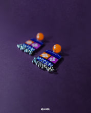 Low-angle view of the Leheriya traditional art drop earrings, showcasing bright orange round studs attached to blue rectangular drops with orange and magenta blocks, detailed with white geometric patterns and dotted accents, finished with dangling silver beads, emphasizing their colorful and artistic craftsmanship – Muselot.