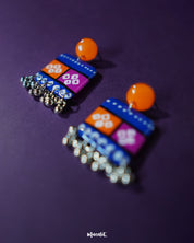 Zoomed-in view of the Leheriya traditional art drop earrings, featuring bright orange round studs connected to blue rectangular drops with orange and magenta blocks, adorned with white geometric patterns and dotted detailing, finished with dangling silver beads, highlighting their intricate handcrafted design – Muselot.