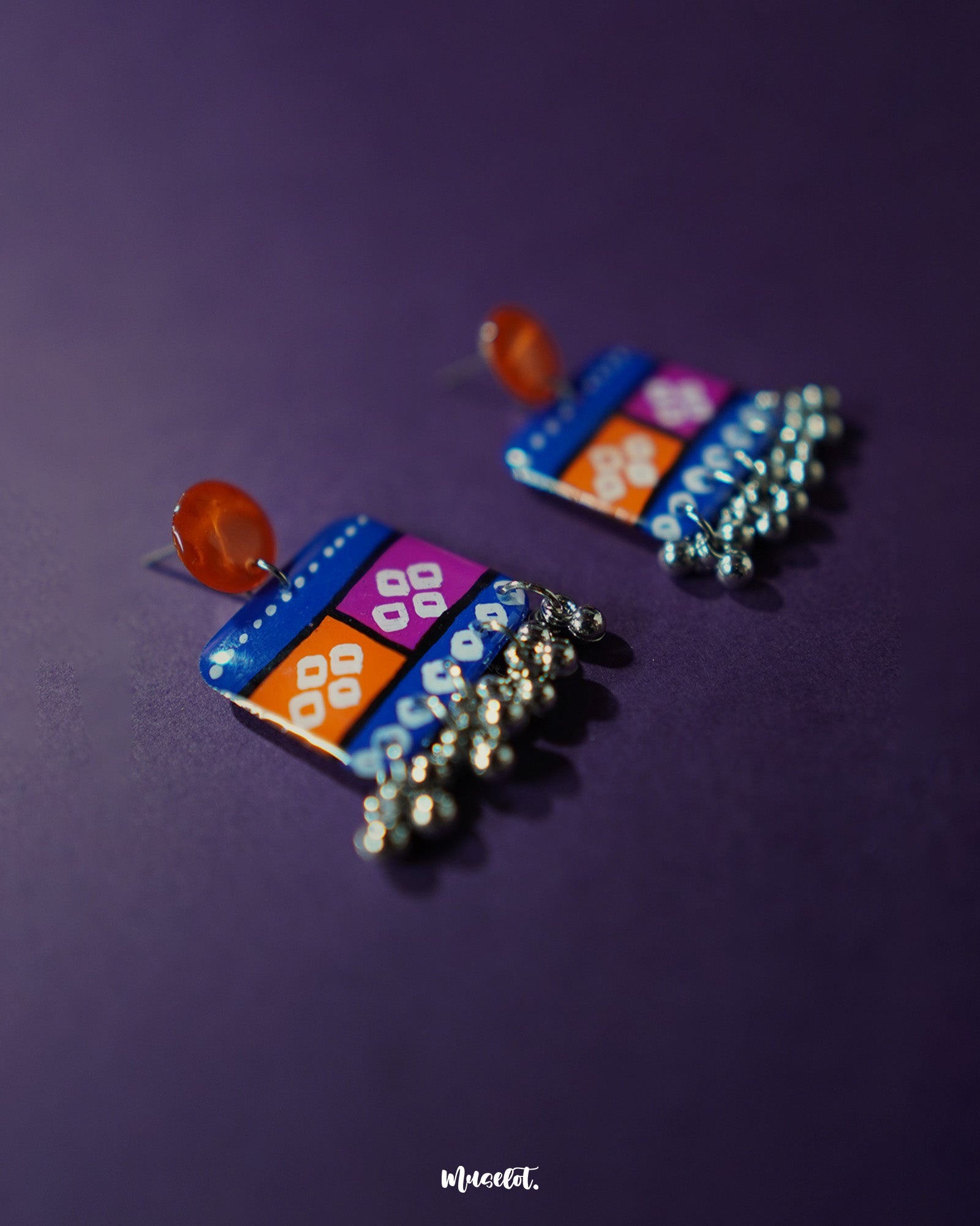 Side view of the Leheriya traditional art drop earrings, showcasing bright orange round studs attached to blue rectangular drops with orange and magenta blocks, detailed with white geometric patterns and dotted accents, finished with dangling silver beads, emphasizing their vibrant and unique design – Muselot.