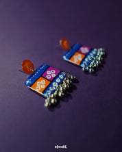 Side view of the Leheriya traditional art drop earrings, showcasing bright orange round studs attached to blue rectangular drops with orange and magenta blocks, detailed with white geometric patterns and dotted accents, finished with dangling silver beads, emphasizing their vibrant and unique design – Muselot.