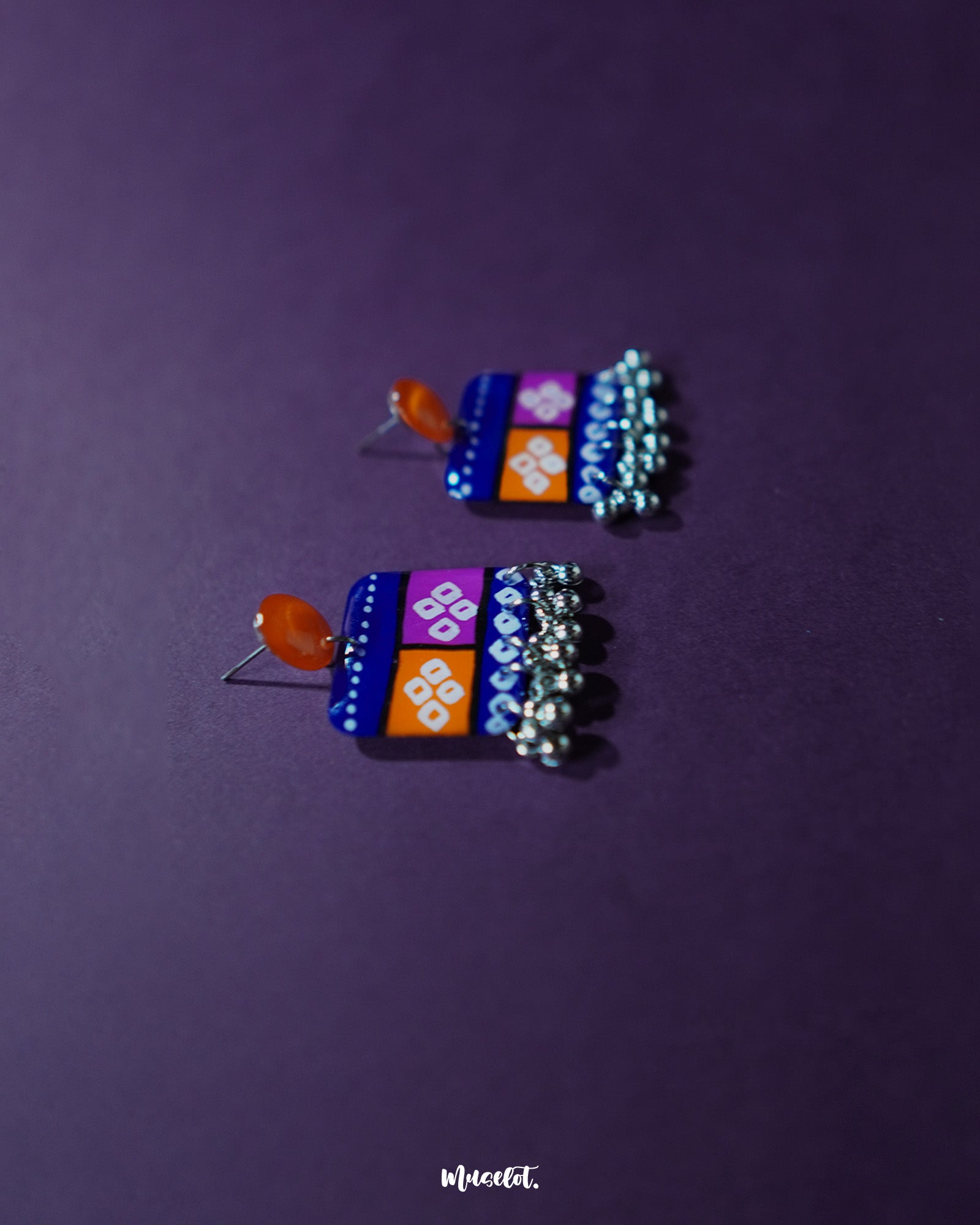 Top side view of the Leheriya traditional art drop earrings, featuring bright orange round studs connected to blue rectangular drops with orange and magenta blocks, adorned with white geometric patterns and dotted detailing, finished with dangling silver beads, capturing their colorful and artistic appeal – Muselot.
