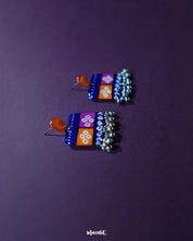 Top side view of the Leheriya traditional art drop earrings, featuring bright orange round studs connected to blue rectangular drops with orange and magenta blocks, adorned with white geometric patterns and dotted detailing, finished with dangling silver beads, capturing their colorful and artistic appeal – Muselot.