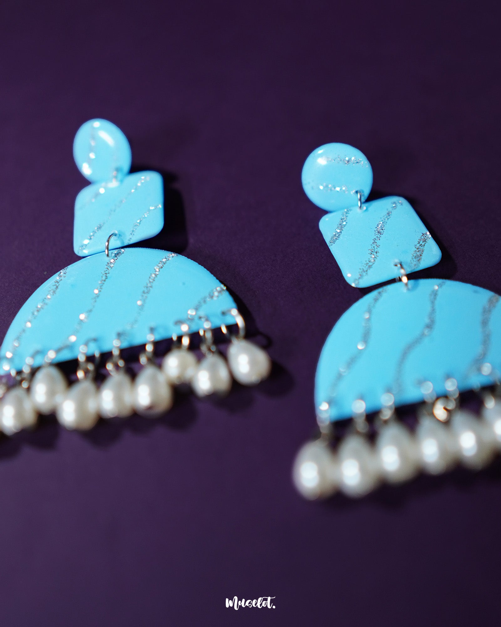 The Lehrbaan blue pearl jhumkas bring a refreshing twist to traditional design — featuring a sky blue base with sparkling silver accents and finished with a row of dainty pearl drops. Their lightweight charm and bold shape make them a vibrant pick for festive flair or a fusion outfit — Muselot.