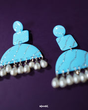 The Lehrbaan blue pearl jhumkas bring a refreshing twist to traditional design — featuring a sky blue base with sparkling silver accents and finished with a row of dainty pearl drops. Their lightweight charm and bold shape make them a vibrant pick for festive flair or a fusion outfit — Muselot.