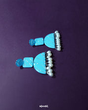 Lehrbaan blue pearl jhumkas resting against a curved neutral backdrop, highlighting the rich enamel finish and cascading pearls with a touch of elegance and depth — Muselot.