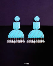 Lehrbaan drop earrings in a striking sky blue hue, detailed with silver glittery waves across geometric shapes. The semicircular base is finished with a row of dangling white pearls, creating a blend of playful charm and elegant contrast — Muselot.