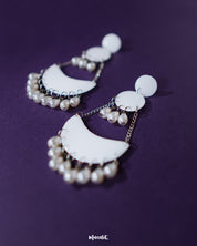 A pair of Mahtab pearl chandbali earrings resting elegantly against a soft backdrop, highlighting the graceful curve of the crescent design adorned with white enamel and hanging pearls — Muselot.