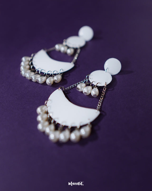 A pair of Mahtab pearl chandbali earrings resting elegantly against a soft backdrop, highlighting the graceful curve of the crescent design adorned with white enamel and hanging pearls — Muselot.