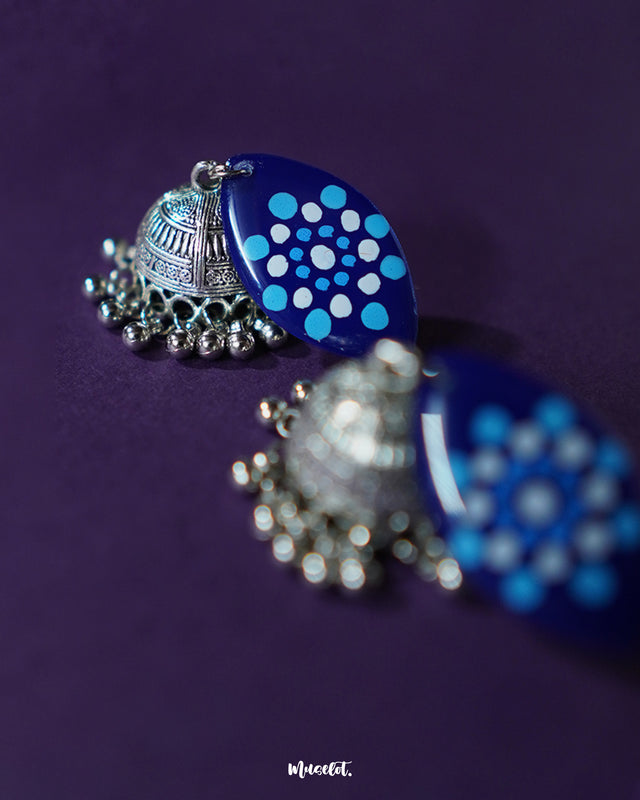 A close-up of Meghmalhar boho jhumkas showing the intricate hand-painted patterns on the blue stud tops and the antique silver domes with delicate ghungroos, highlighting artisanal detailing and showcasing timeless charm — Muselot.