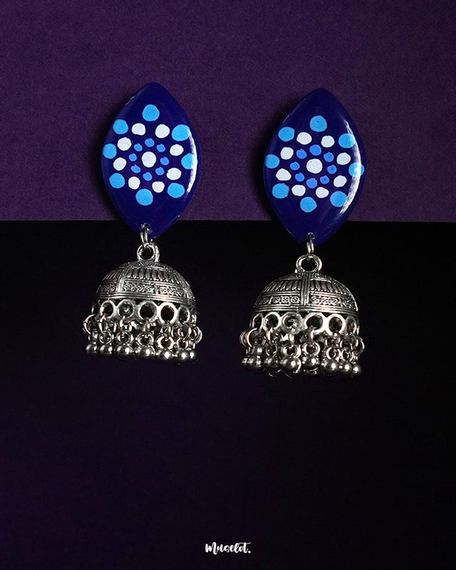 A pair of Meghmalhar jhumkas featuring deep blue marquise-shaped tops with dotted circular sky blue and white patterns, complemented by intricately detailed oxidized silver domes and ghungroo-style danglers, showcasing a striking fusion of tradition and bold design — Muselot.
