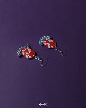 The Mehar red floral hook earrings from Muselot laid flat — highlighting the symmetrical hexagonal shape, vivid red enamel, delicate white floral detailing, and metallic ghungroo accents that showcase the piece’s handcrafted charm and cultural finesse.