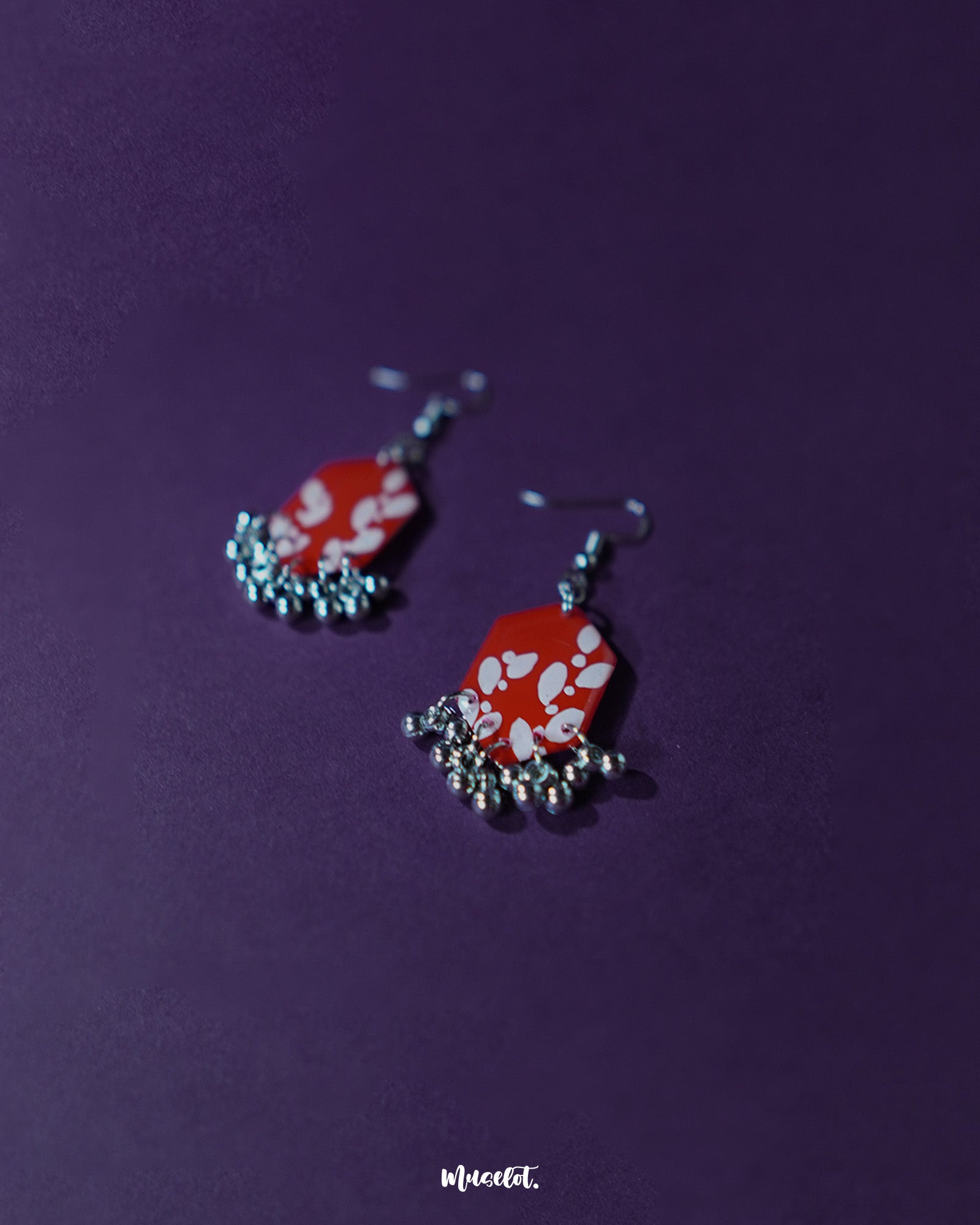The Mehar red floral hook earrings from Muselot arranged elegantly — highlighting their geometric silhouette, rich red tones, and hand-painted white floral patterns, with metallic ghungroos adding a rhythmic charm to the design.