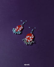 The Mehar red floral hook earrings from Muselot arranged elegantly — highlighting their geometric silhouette, rich red tones, and hand-painted white floral patterns, with metallic ghungroos adding a rhythmic charm to the design.