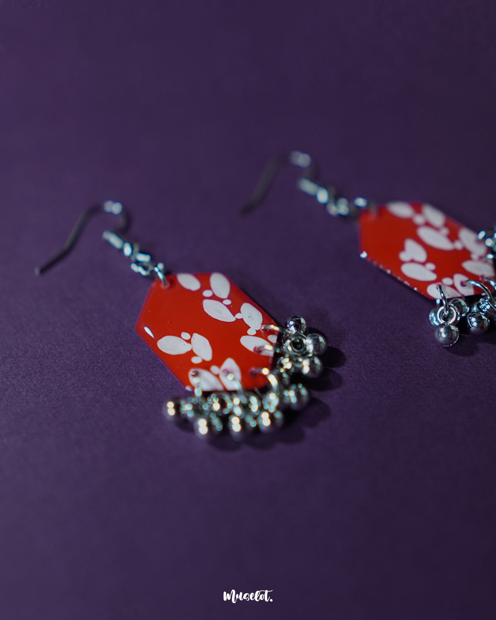 A close-up view of the Mehar red floral hook earrings from Muselot — highlighting the fine brushwork of the white floral motifs on vivid red enamel drops, with metallic ghungroos accentuating the handcrafted detail and traditional artistry.