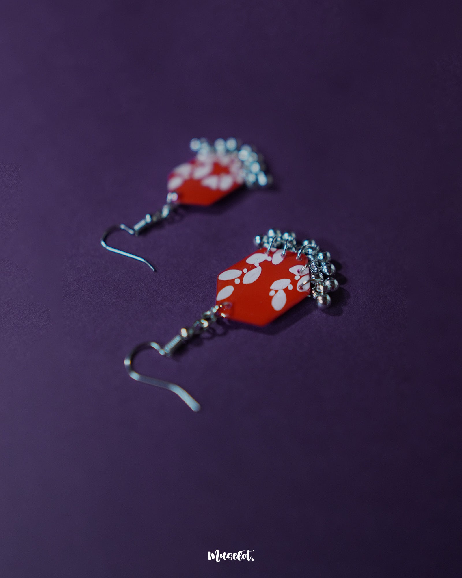 The Mehar red floral hook earrings from Muselot styled with complementary props — showcasing the vibrant red enamel, hand-painted white floral designs, and metallic ghungroos in a visually rich composition that highlights their festive and artisanal appeal.