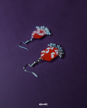 The Mehar red floral hook earrings from Muselot styled with complementary props — showcasing the vibrant red enamel, hand-painted white floral designs, and metallic ghungroos in a visually rich composition that highlights their festive and artisanal appeal.