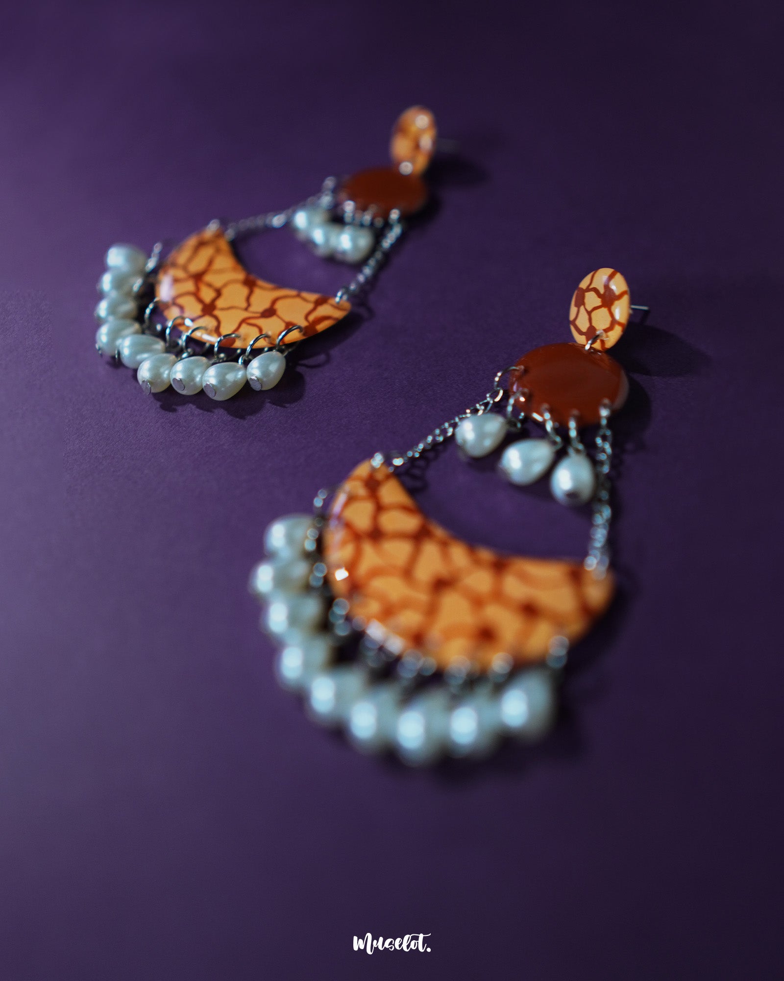 A close-up of the Mittika chandbali earrings placed on a soft brown fabric, showcasing their handcrafted orange enamel finish, delicate pearl detailing, and oxidized silver textures that blend rustic charm with festive elegance — Muselot.