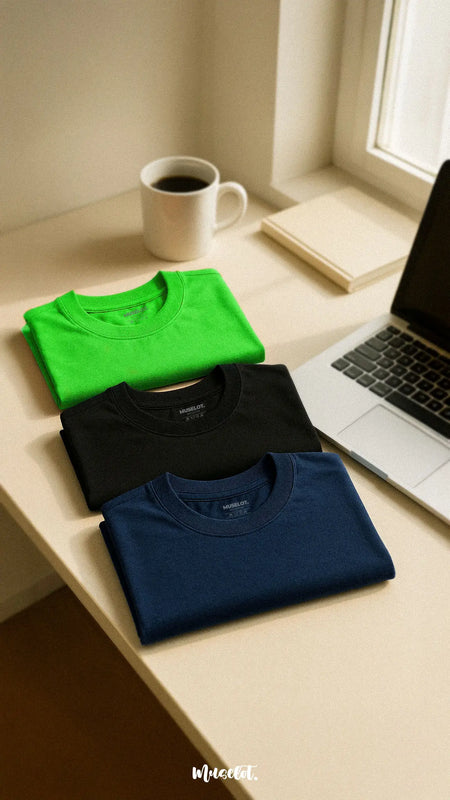 A cozy set of soft cotton T-shirts in lilac green, black, and navy blue, beautifully laid out on a warm wooden surface, showcasing their soothing colors and relaxed fit. These tones evoke a lounge vibe, perfect for coffee breaks and chill days — Muselot.