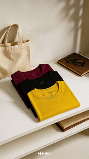 A cozy combo of soft cotton T-shirts in mustard yellow, deep black, and rich maroon, laid out side by side on a warm, linen-textured backdrop. The relaxed silhouettes and muted tones evoke a bookish calm and laid-back lounge aesthetic, making them perfect for slow days, warm tea, and quiet reads — Muselot