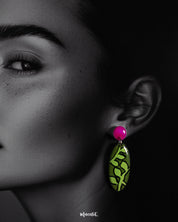 A woman wears the Naghma handmade leaf drop earrings, captured in a close-up that highlights the vibrant green leaf pattern and glossy magenta studs, showcasing the hand-painted detailing and bold botanical charm — Muselot.
