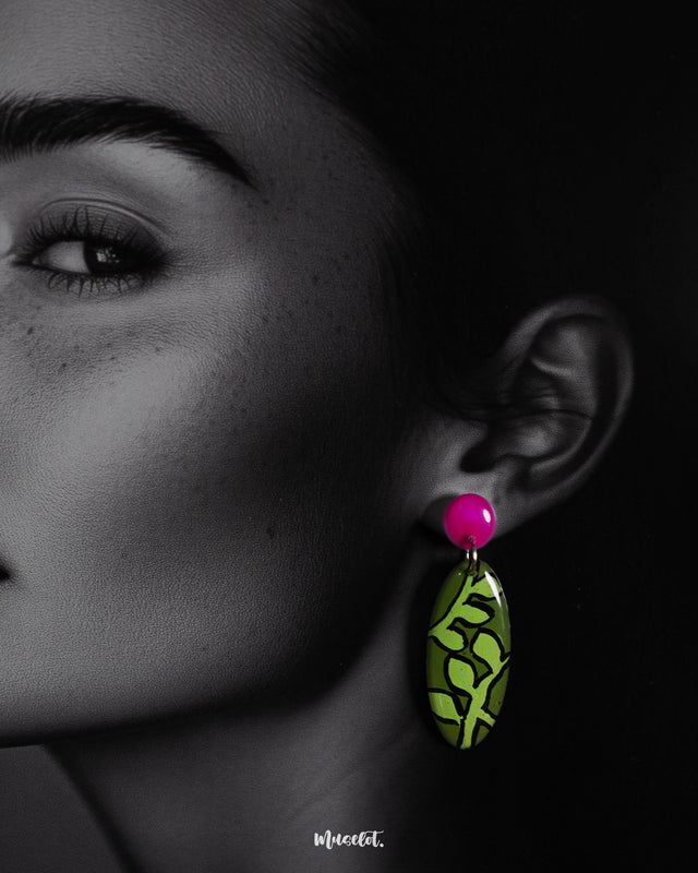 A woman wears the Naghma handmade leaf drop earrings, captured in a close-up that highlights the vibrant green leaf pattern and glossy magenta studs, showcasing the hand-painted detailing and bold botanical charm — Muselot.