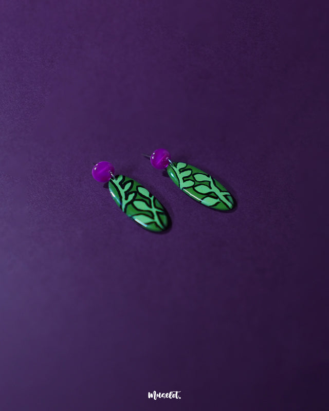 A pair of Naghma green leaf drop earrings featuring a vibrant green base with bold black leaf patterns and glossy magenta studs, neatly displayed against a dual-toned purple backdrop, highlighting and showcasing their artistic, hand-painted charm — Muselot.