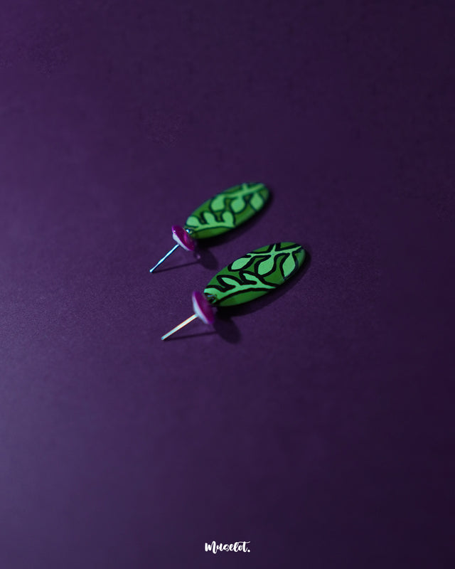 A close-up view of the Naghma green leaf drop earrings showcasing their sleek, oval silhouette with detailed black leaf motifs on a vibrant green base, paired with glossy magenta studs for a playful contrast — Muselot.