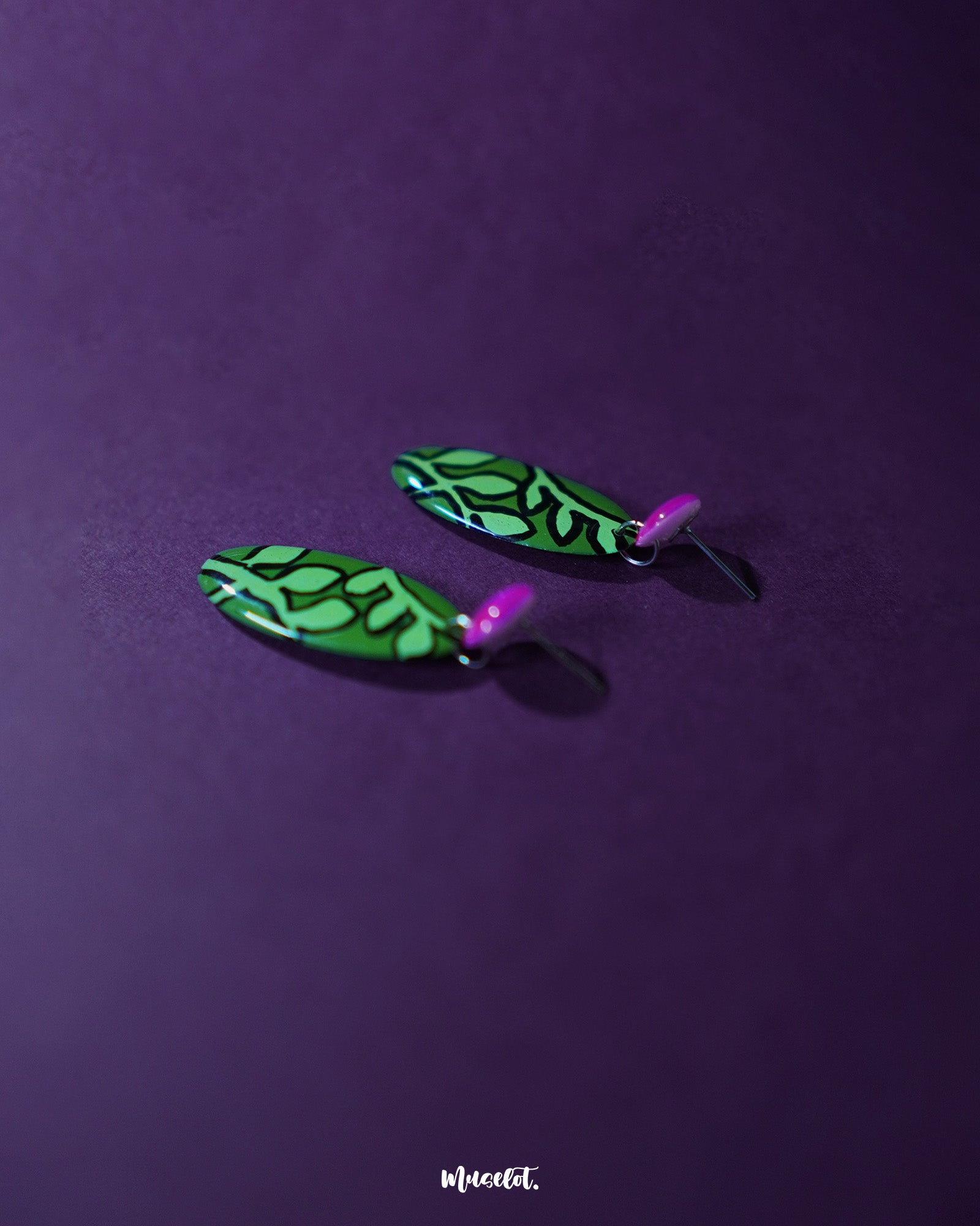 A pair of Naghma green leaf drop earrings displayed against a purple background, highlighting the striking hand-painted leaf pattern in black over a vivid green base, attached to glossy magenta studs that enhance their bold, artistic appeal — Muselot.