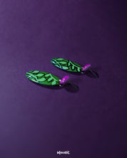 A pair of Naghma green leaf drop earrings displayed against a purple background, highlighting the striking hand-painted leaf pattern in black over a vivid green base, attached to glossy magenta studs that enhance their bold, artistic appeal — Muselot.