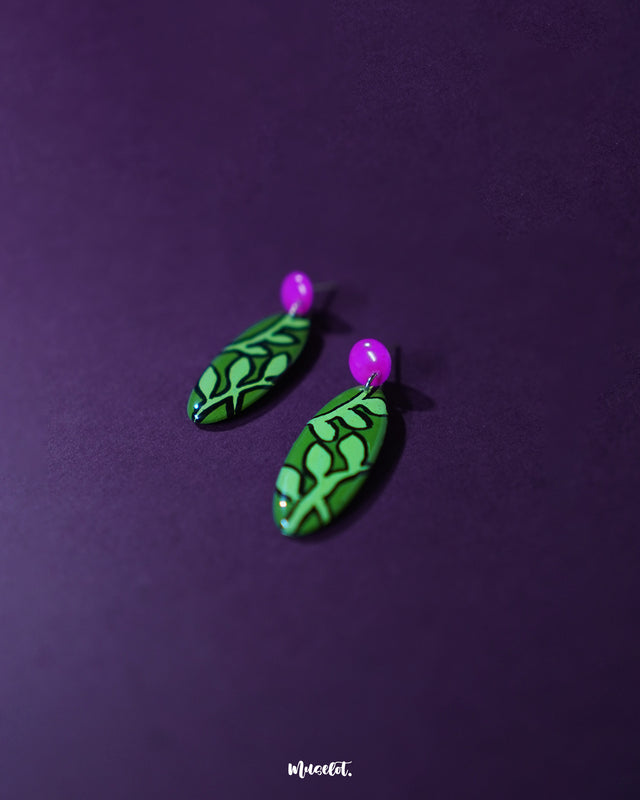 Naghma green leaf drop earrings shown flat against a deep purple backdrop, showcasing their elongated oval shape, black-outlined leaf motifs on a bright green surface, and the glossy pink studs that add a vibrant contrast — Muselot.