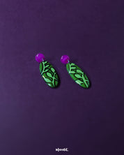 A close-up view of the Naghma green leaf drop earrings, highlighting the intricate veined leaf pattern outlined in black, the rich green enamel finish, and the soft gloss of the pink studs for a striking contrast — Muselot.