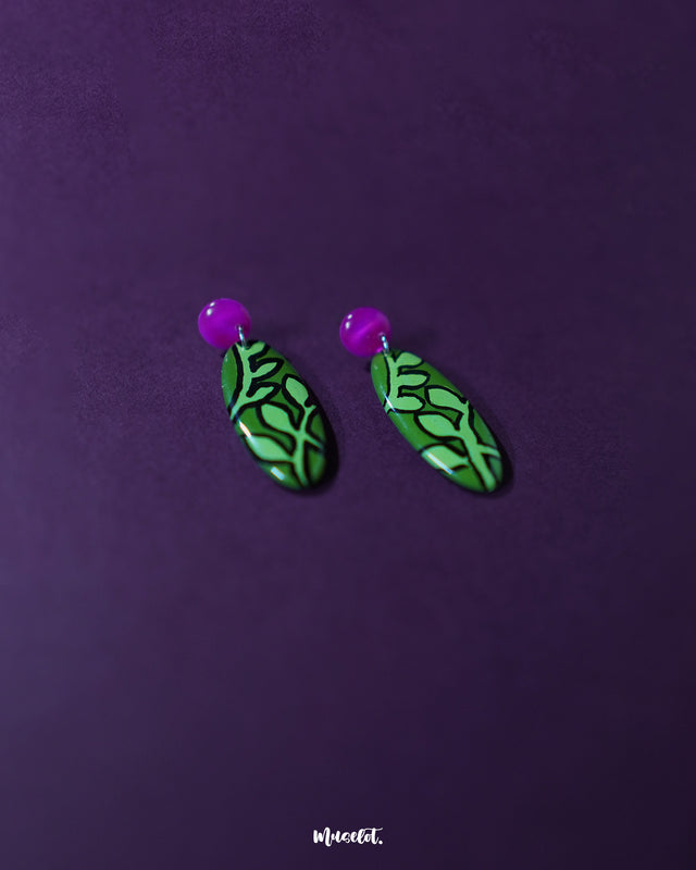 A close-up view of the Naghma green leaf drop earrings, highlighting the intricate veined leaf pattern outlined in black, the rich green enamel finish, and the soft gloss of the pink studs for a striking contrast — Muselot.