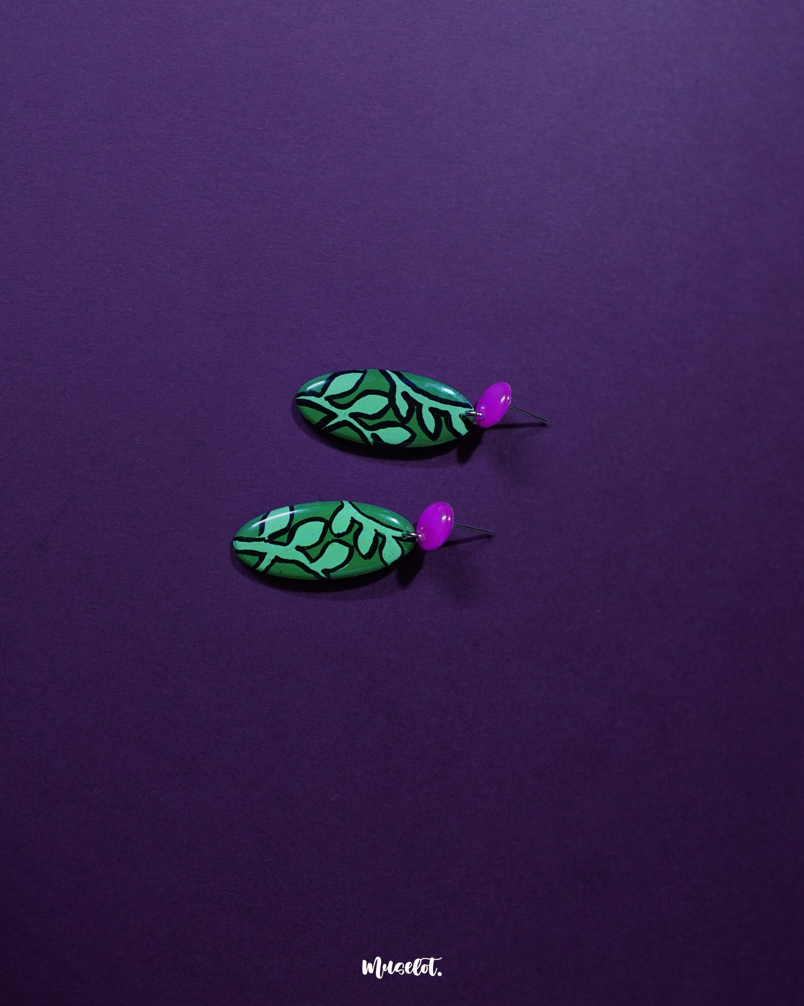 A detailed view of the Naghma green leaf drop earrings laid out flat, highlighting the bold green enamel, delicate black veining, and pink stud tops that bring a refreshing, nature-inspired charm — Muselot.