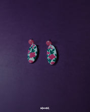 Nargis pink floral drop earrings displayed against a purple background, highlighting the hand-painted rose pink floral motifs and glossy finish that showcase their playful charm and artisanal elegance — Muselot.