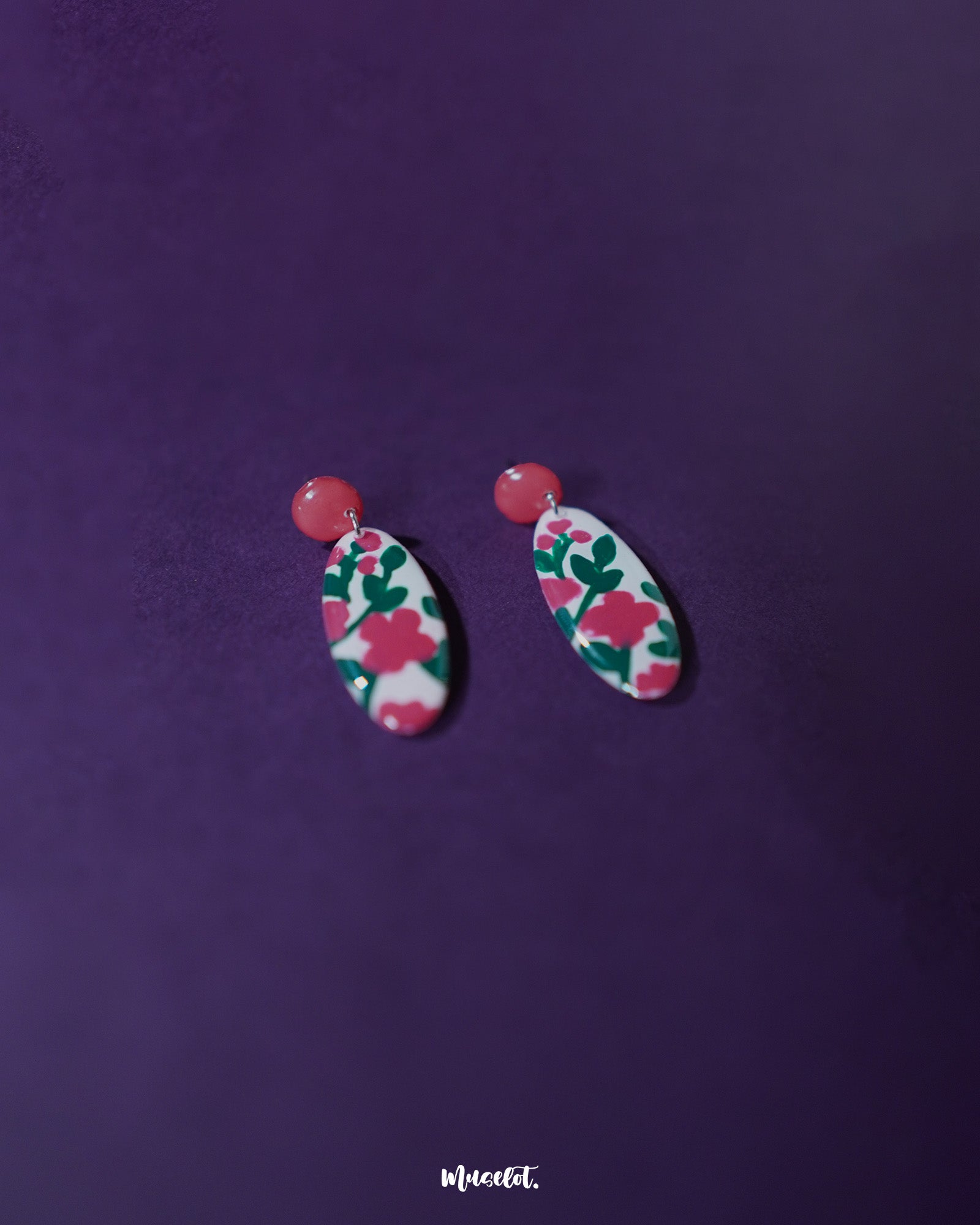 A pair of Nargis pink floral drop earrings resting flat, showcasing the oval-shaped design with vivid pink flowers and green leaves on a white base, highlighting the glossy enamel and handmade details — Muselot.