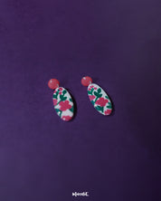 A pair of Nargis pink floral drop earrings resting flat, showcasing the oval-shaped design with vivid pink flowers and green leaves on a white base, highlighting the glossy enamel and handmade details — Muselot.