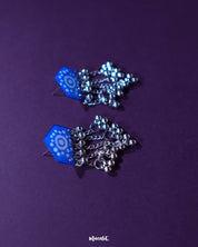 Nazafar blue jhumkas with white hand-painted motifs and silver dome drops, displayed against a purple background to highlight their bold traditional design – Muselot.