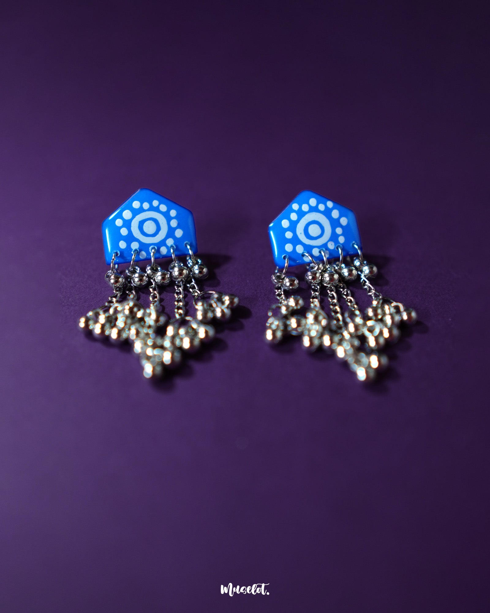 Pair of Nazafar blue jhumkas displayed flat on a neutral surface, showcasing bold hand-painted motifs while highlighting the traditional silver dome and folk-inspired craftsmanship – Muselot.