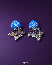 Pair of Nazafar blue jhumkas displayed flat on a neutral surface, showcasing bold hand-painted motifs while highlighting the traditional silver dome and folk-inspired craftsmanship – Muselot.